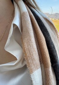 Cachecol com riscas alternadas em bege, castanho claro e cinza escuro, feito de um tecido de lã suave e texturizado, colocado sobre uma camisa branca.