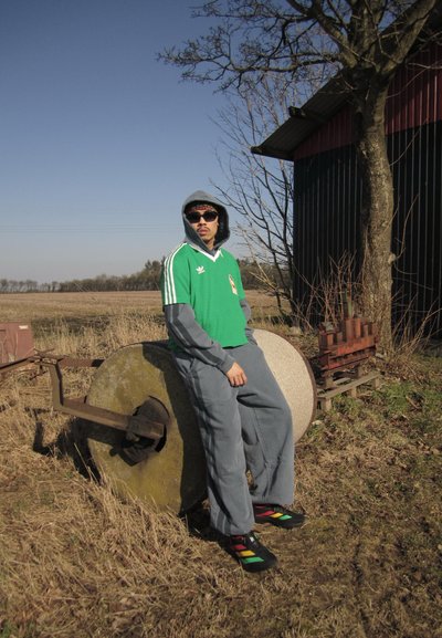 Hombre con camiseta verde de Adidas, sudadera gris y zapatos coloridos apoyado contra un rodillo agrícola grande cerca de un granero y un árbol en un campo seco bajo un cielo despejado.