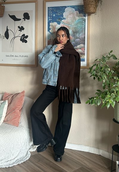 Chaqueta de mezclilla, bufanda marrón con flecos, pantalones de campana negros y zapatos negros gruesos. Entorno interior con arte, plantas y cojines.