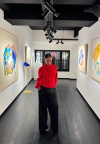 Czerwony sweter w prążki z guzikami na ramionach, czarne szerokie spodnie oraz sandały na odkryte palce, w nowoczesnej galerii sztuki z kolorowymi obrazami.