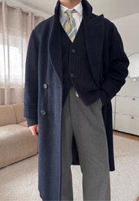 Homme portant un pardessus bleu marine, un cardigan noir, une chemise blanche, une cravate rayée et un pantalon gris, debout dans un salon avec un canapé beige et une armoire.