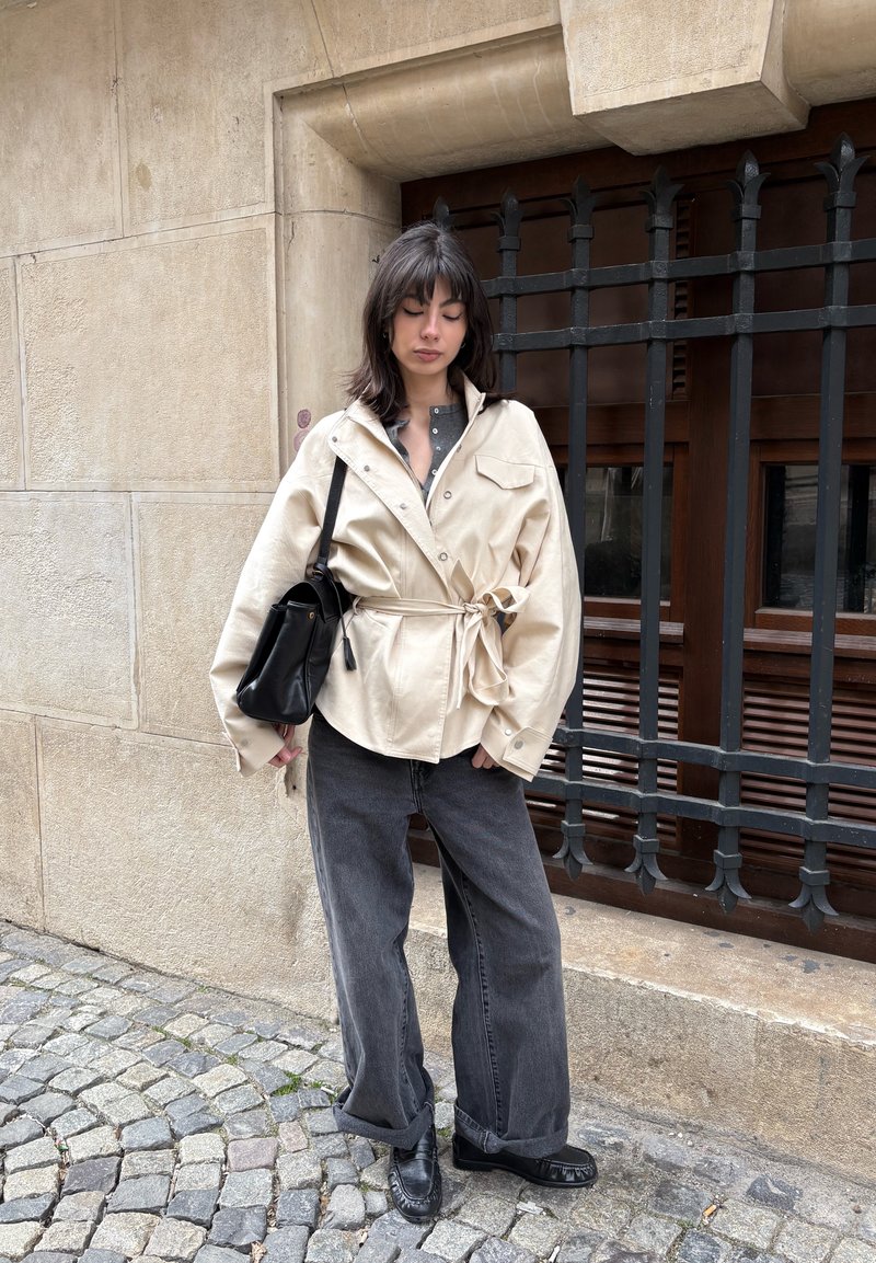 Femme debout dans une rue pavée portant une veste beige oversize nouée à la taille, un jean foncé ample, des chaussures noires, et portant un sac à bandoulière noir.