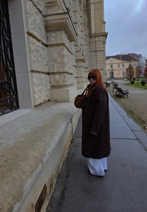 Sieviete ar sarkaniem matiem un saulesbrillēm stāv uz mitra bruģa blakus reljefai akmens ēkai zem mākoņainas debess, nesot brūnu somu.