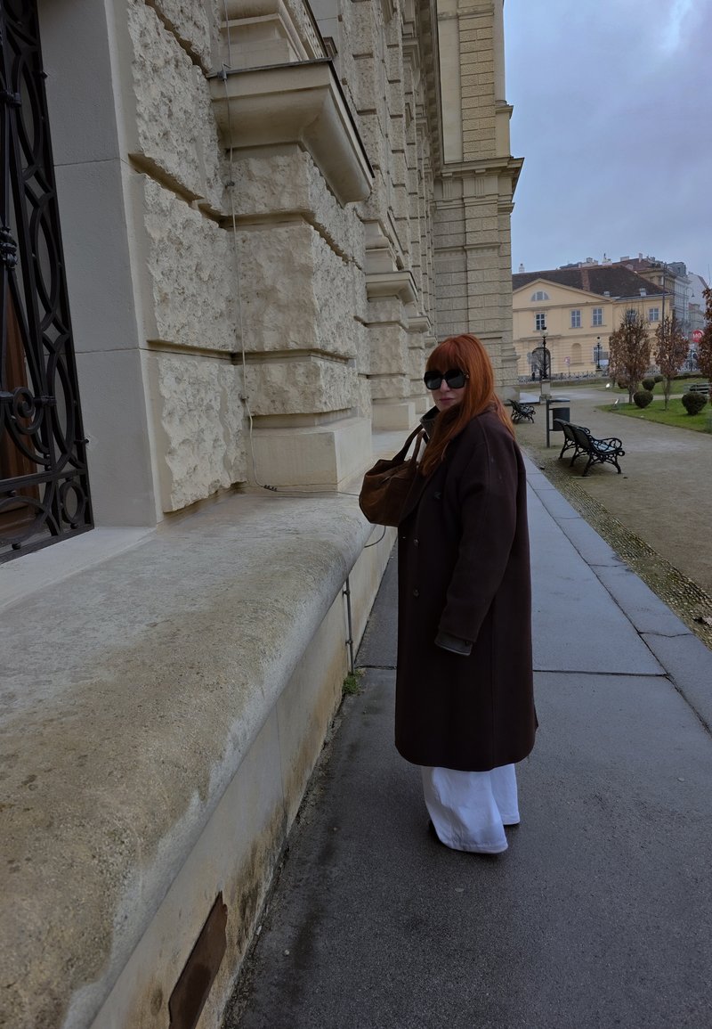 Γυναίκα με κόκκινα μαλλιά και γυαλιά ηλίου στέκεται σε βρεγμένο πεζοδρόμιο δίπλα σε κτήρια με υφή πέτρας κάτω από συννεφιασμένο ουρανό, κρατώντας καφέ τσάντα.