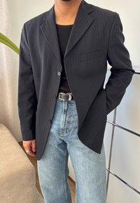 Blazer preto risca de giz, com um bolso, três botões, usado sobre uma camisola preta de alças, combinado com jeans largos de um azul claro. Design simples e bem ajustado.