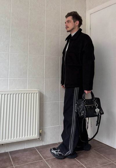 Chaqueta negra con cremallera, camiseta gris, jeans negros con franjas laterales, zapatillas negras y un bolso pequeño con detalles en un ambiente de habitación con azulejos.