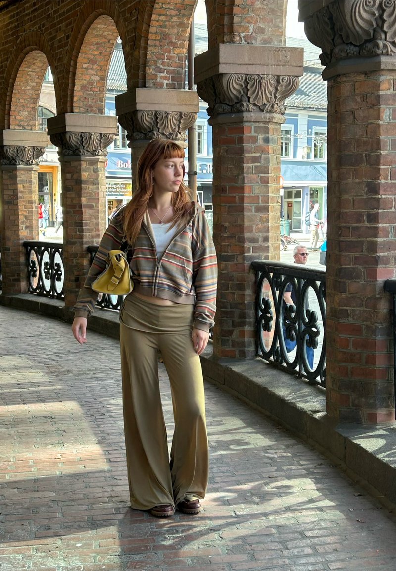 Jeune femme portant une veste rayée et un sac jaune, debout sous des arcades en briques sur un passage ensoleillé, regardant sur le côté.