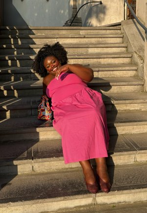 Vestido maxi rosa sin mangas con cintura fruncida, combinado con tacones burdeos. Un bolso de patrón colorido yace al lado en los escalones de concreto.