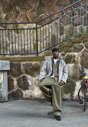 Chaqueta gris de gran tamaño sobre un conjunto verde olivo, sentada contra una pared de piedra con un pasamanos negro y una bicicleta cerca.