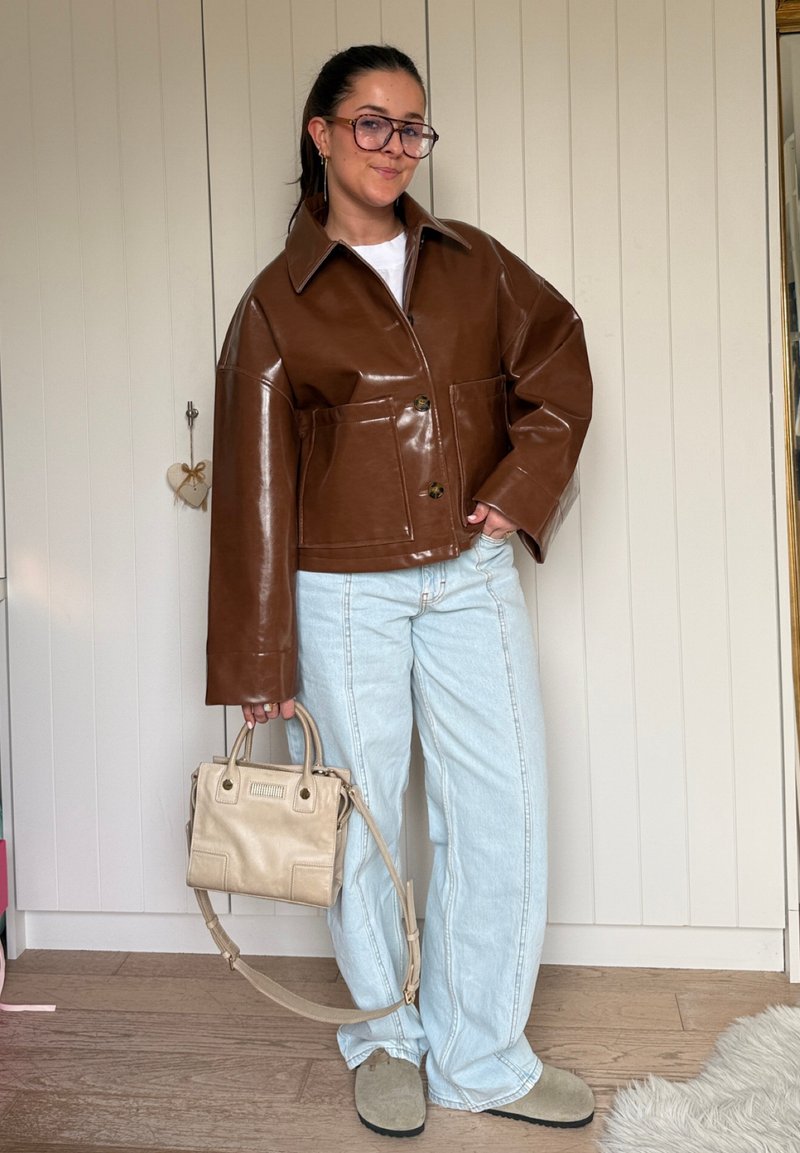 Woman wearing brown leather jacket, light blue jeans, beige shoes, holding beige handbag, standing in a room with wooden floor and white paneled wall.
