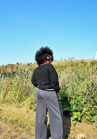 Melna adīta džemperis ar baltām akcentiem, pelēkas platās bikses, čirkainas matu frizūras, stāvot zālē zem skaidras zilas debess.