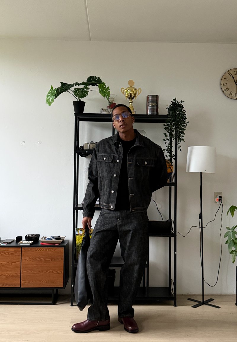 Homme portant une veste et un pantalon en denim foncé, tenant un sac noir, debout dans un salon avec des plantes, une horloge, une étagère et une lampe sur pied.