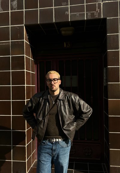 Hombre con una chaqueta de cuero negro sobre un suéter oscuro, jeans de mezclilla claros y un cinturón estampado, de pie junto a una pared de azulejos oscuros con una puerta roja.