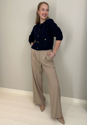 Cárdigan de punto con cable en azul marino con botones dorados, pantalones de cintura alta en color crema y tacones beige de punta afilada, frente a una pared de color claro.