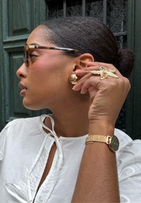 Occhiali da sole tartarugati, orecchini rotondi dorati, un orologio dorato con texture e un anello d'oro unico a forma di stella marina. Blusa bianca con ricami.