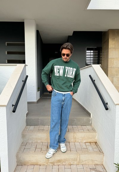 Sudadera verde con el texto "NUEVA YORK", jeans sueltos azules y zapatillas blancas. De pie en los escalones, usa gafas de sol y tiene el pelo de longitud media.