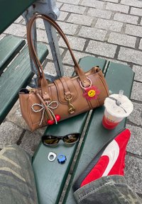 Brun håndveske med gullhardware, nøkkelelementer, solbriller, fargerike ringer, og røde semsket skinnsko på en grønn benk med steinbelagt fortau.