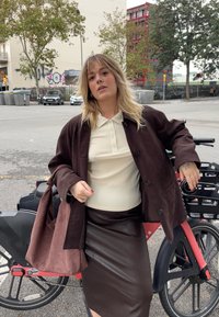 Dunkelbraune Jacke über einem hellen Poloshirt, kombiniert mit einem dunkelbraunen Lederrock. Eine pinke Tasche und ein Fahrrad im Hintergrund. Natürliche Outdoor-Umgebung.