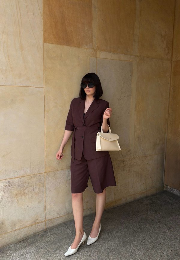Femme avec une coupe carré foncée portant des lunettes de soleil surdimensionnées, un blazer marron foncé et un short, des talons blancs, tenant un sac à main beige, debout contre un mur de pierre.