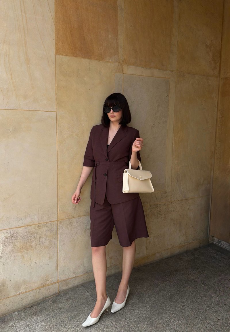 Femme avec une coupe carré foncée portant des lunettes de soleil surdimensionnées, un blazer marron foncé et un short, des talons blancs, tenant un sac à main beige, debout contre un mur de pierre.