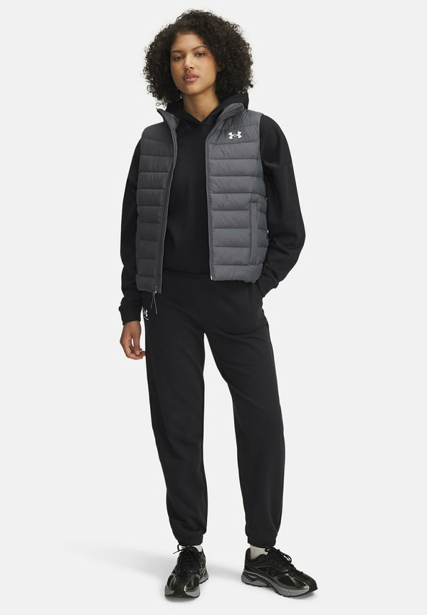 Grijze puffer vest met ritssluiting, voorzien van horizontale stiksels, gedragen over een zwart hoodie en zwarte trainingsbroek, aangevuld met zwarte schoenen.