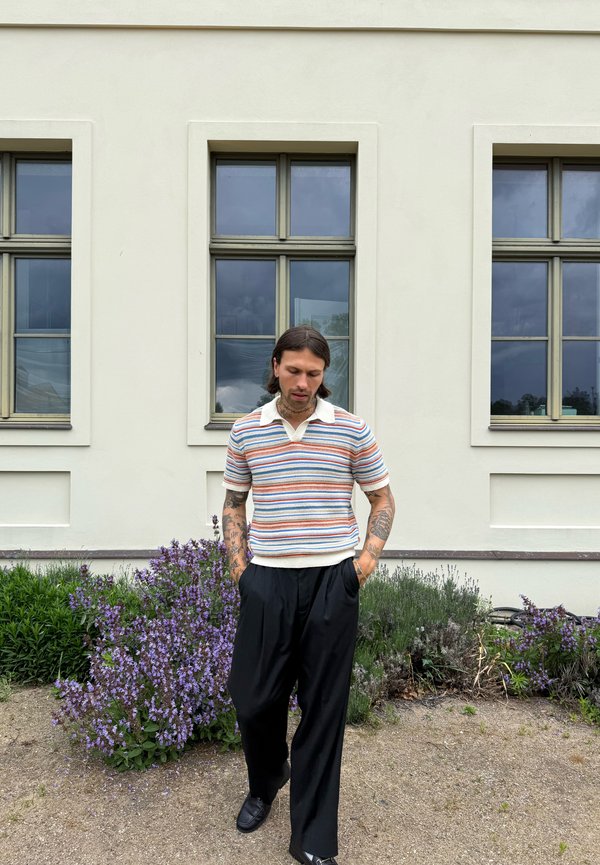 Homme tatoué portant une chemise rayée et un pantalon noir se tient près de fleurs violettes devant un bâtiment beige avec trois fenêtres.