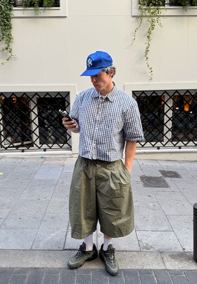 Conjunto casual que incluye una camisa de cuadros de manga corta, pantalones cortos sueltos de color verde oliva, calcetines blancos y zapatillas oscuras, complementado con una gorra azul.