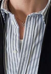 Collier en argent avec un pendentif circulaire ; chemise à col à rayures verticales bleues et blanches ; boutons visibles ; détail de veste noire.