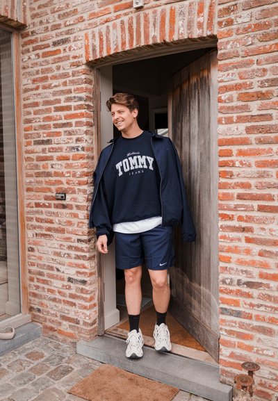 Sudadera marinera con el texto "TOMMY", pantalones cortos a juego y zapatillas blancas. De pie junto a una puerta con un marco de madera rústica y pared de ladrillo.
