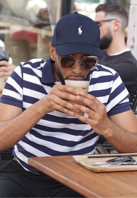 Camisa polo a rayas en azul marino y blanco, gorra azul marino, gafas de sol grandes, sosteniendo un vaso transparente de café helado, sentado en una mesa de madera con utensilios.