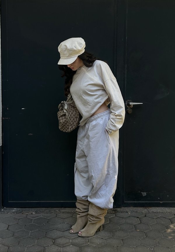 Kvinde i beige outfit med kortærmet top, løstsiddende bukser, rummelige hælede støvler og kasket står og kigger ned mod en mørk dør.