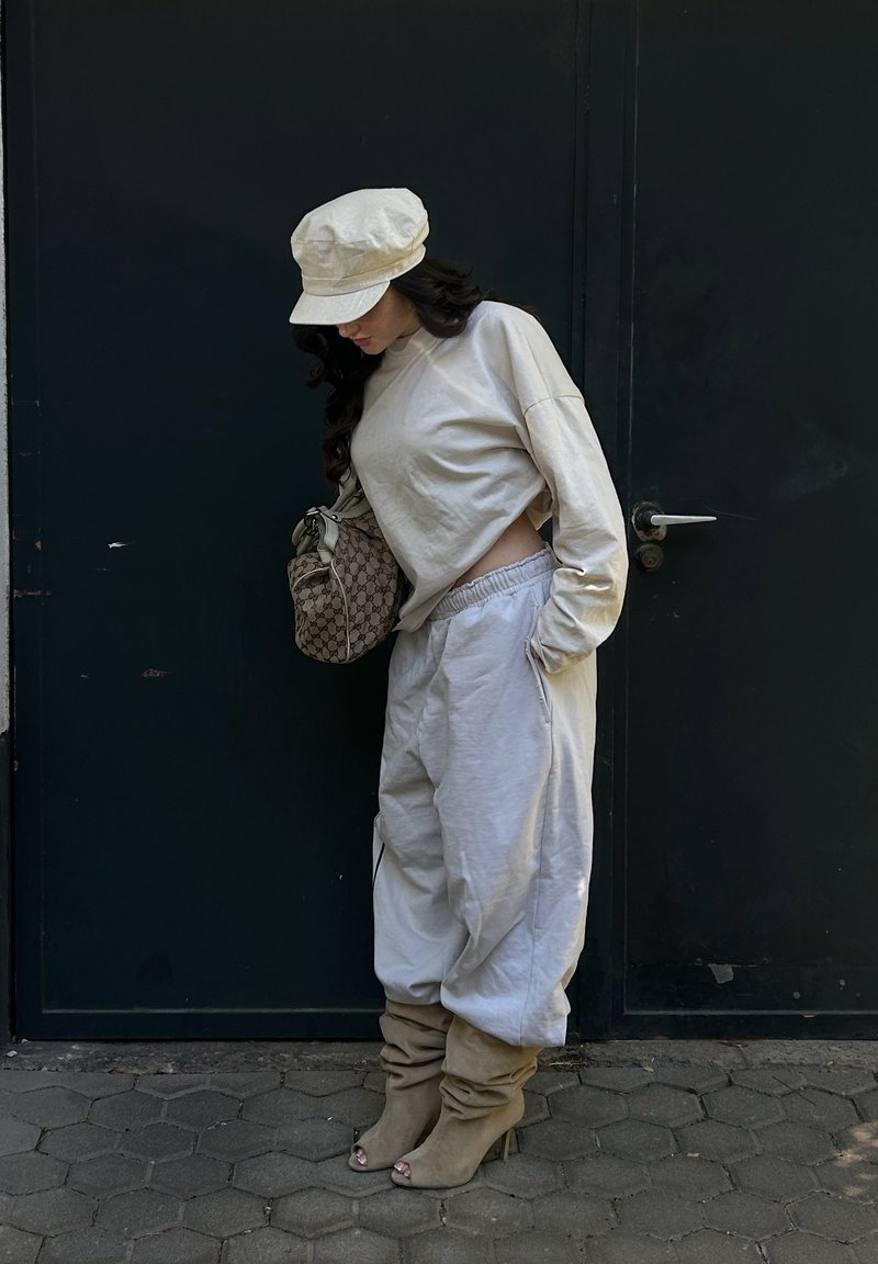 Mujer con conjunto beige con top de manga larga recortado, pantalones anchos, botas con tacón y caída casual, y gorra, está de pie mirando hacia abajo junto a una puerta oscura.