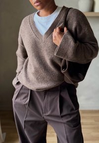 Άτομο που φοράει μπεζ πουλόβερ με V λαιμόκοψη πάνω από λευκό πουκάμισο, σκούρο πλισέ παντελόνι και κρατά μια σκούρα καφέ τσάντα στον ώμο.