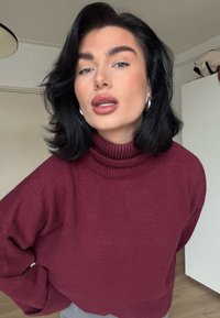 Pull col roulé marron avec col côtelé, texture douce et coupe ample. Présente des cheveux noirs ondulés et des boucles d'oreilles en créole sur un fond neutre.