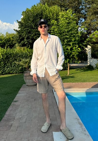 Camisa de lino blanca ligera, pantalones cortos rayados beige y zapatos slip-on grises, situados junto a una piscina rodeada de follaje verde y pavimento de piedra.