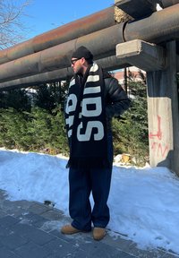Man in een zwarte beanie, zonnebril, leren jas, brede broek en een grote zwart-wit sjaal staat op een besneeuwd trottoir naast roestige buizen.
