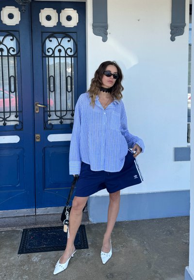 Camisa de rayas azules, de corte holgado, abotonada, combinada con pantalones cortos navy con rayas blancas en los laterales. Mulas blancas con tacón y un bolso negro complementan el conjunto.