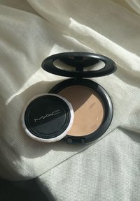 Schwarze Kompaktpuderbox geöffnet, die ein hellbraunes Puder und einen weißen runden Applikator mit schwarzem Rand zeigt. Der Hintergrund hat eine sanfte Textur.