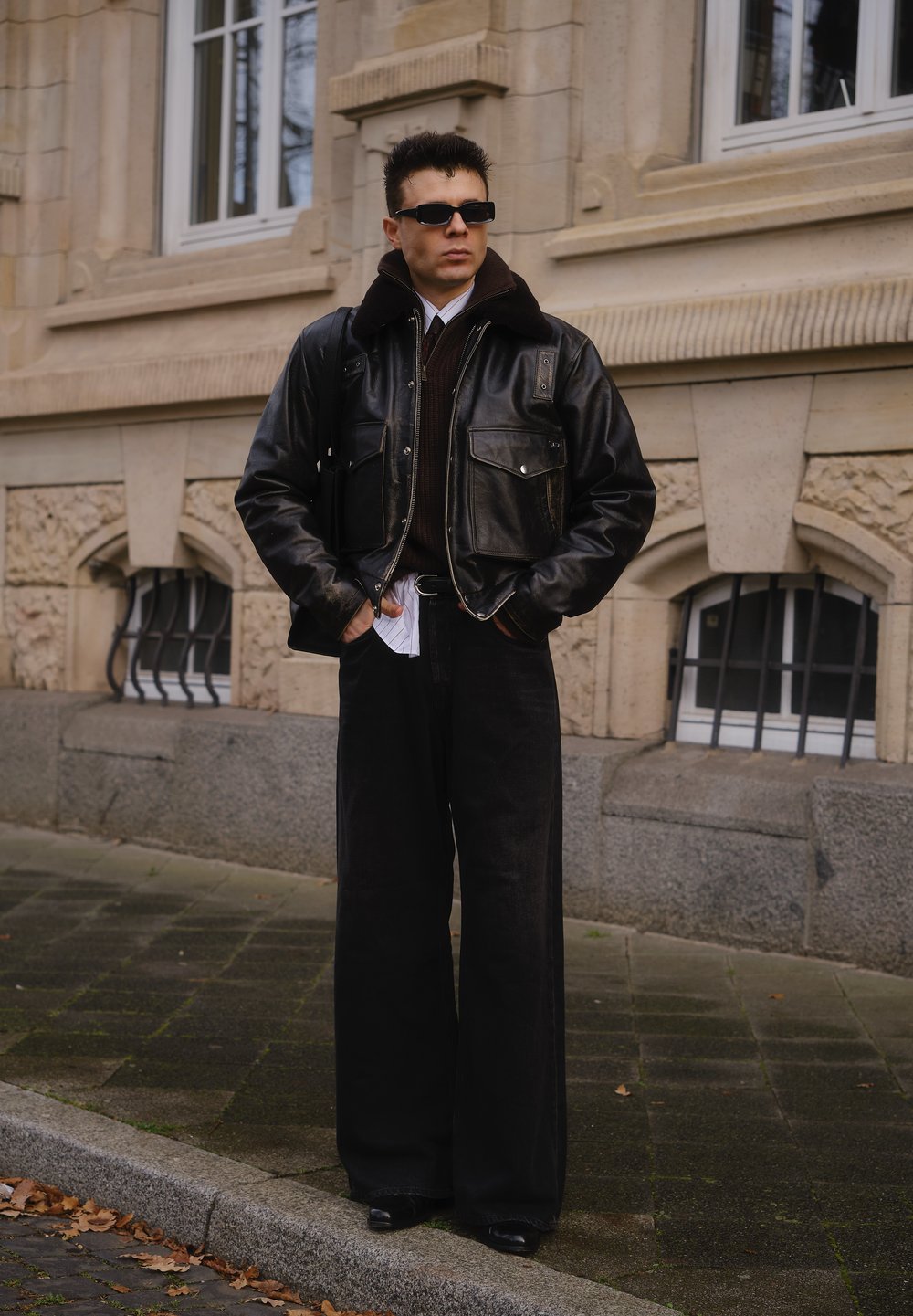 Sort læderbomberjakke med pelskant, mørkebrun sweater, sorte bukser med brede ben og sorte sko. Står udendørs tæt på arkitektur.