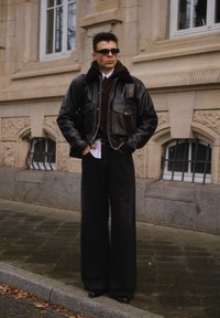 Sort læderbomberjakke med pelskant, mørkebrun sweater, sorte bukser med brede ben og sorte sko. Står udendørs tæt på arkitektur.