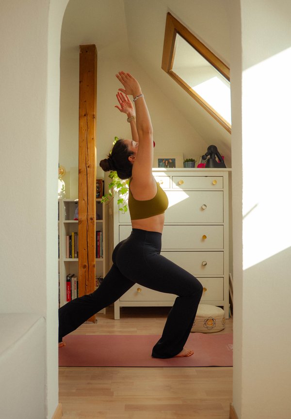 Femme en tenue de sport effectuant une posture du guerrier en yoga sur un tapis dans une pièce baignée de lumière avec une commode, des plantes et des livres.