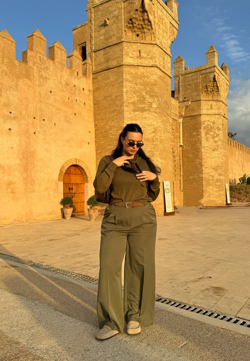 Donna in abito verde oliva che sistema una sciarpa, in piedi davanti a una grande fortezza storica beige con torri e porta ad arco in legno sotto la luce del sole.