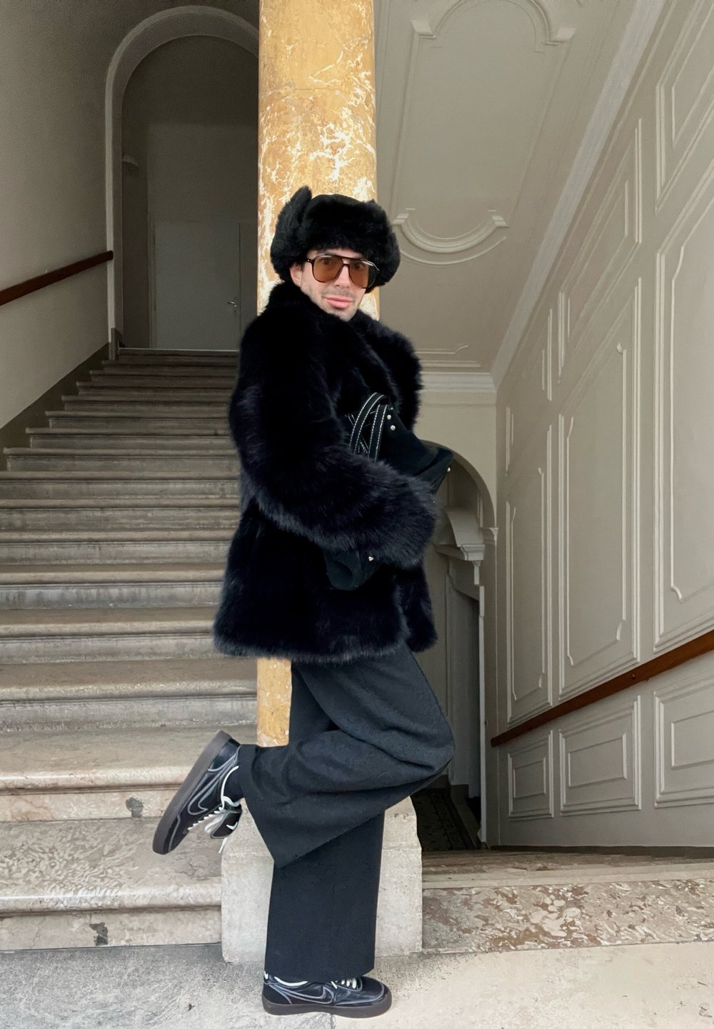 Černý kabát z umělé kožešiny s širokými rukávy, položený přes černý outfit. Osoba má na sobě černou kožešinovou čepici a vzorované boty, pózující proti schodišti.