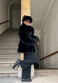 Černý kabát z umělé kožešiny s širokými rukávy, položený přes černý outfit. Osoba má na sobě černou kožešinovou čepici a vzorované boty, pózující proti schodišti.