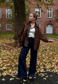 Smeđa suedjana jakna s preklopnim zatvaračima, složena preko karirane košulje i bijele majice. Tamnoplave flare traperice i bež čizme do gležnja upotpunjuju outfit.