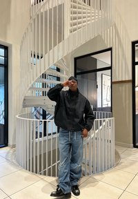 Mann i svart jakke og jeans som justerer brillene, står innendørs foran en hvit spiraltrapp med store vinduer og flislagt gulv.