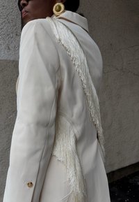 Γυναίκα με εκρού σακάκι με μακριά κρόσσια στους ώμους και στην πλάτη, φοράει μεγάλα στρογγυλά χρυσά σκουλαρίκια και μαύρα γυαλιά ηλίου.