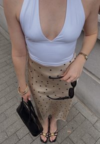 Hvit halterneck topp, beige midi-skjørt med prikker, gull armbånd, svart veske, svarte solbriller, og flate svarte sandaler med gulldetaljer.