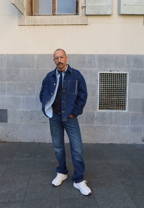 Man with mustache wearing glasses, blue denim jacket, jeans, and white sneakers standing against a tiled wall with a vent.