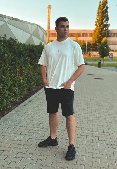 Camiseta blanca de Jordan, pantalones cortos negros desgastados y zapatillas negras en un camino pavimentado rodeado de vegetación y edificios modernos.